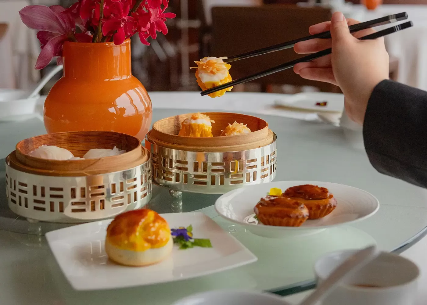 Person with chopsticks, eatting from a variety of dim sum in plates and bamboo bowls at Lung King Heen, Hong Kong.