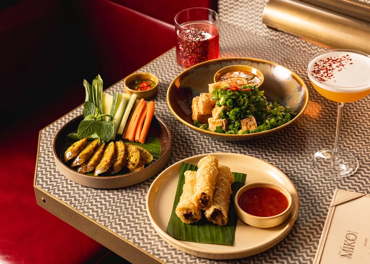 A table with plates of grilled skewers and vegetables, crispy spring rolls with dipping sauce, a salad with fried tofu, and two cocktails, set on a geometric-patterned surface.