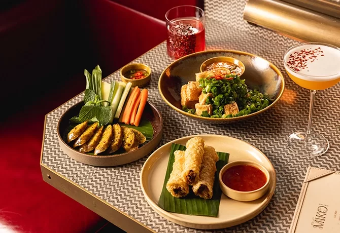 A table set with Vietnamese dishes: grilled meat skewers with fresh vegetables, crispy spring rolls with dipping sauce, a salad with fried toppings, and two colorful drinks. A menu rests beside the plates.