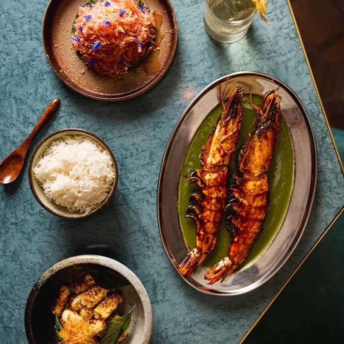 A blue table set with grilled prawns on a green sauce, a bowl of white rice, a plate of salad with shredded toppings, a small bowl of vegetables, and a glass of water.