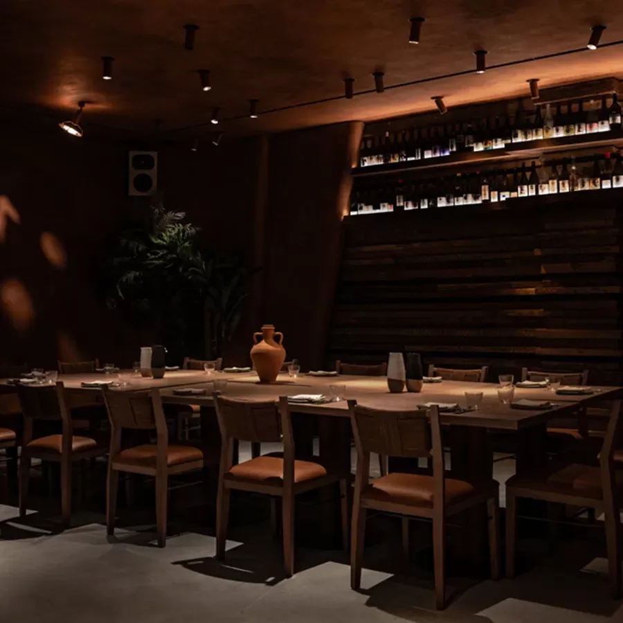 A dimly lit restaurant with a long wooden table, surrounded by wooden chairs. Shelves with bottles line the angled back wall, and a large clay vase is placed at the center of the table. Warm, ambient lighting creates a cozy atmosphere.