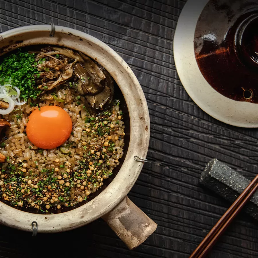 A bowl of rice topped with a raw egg yolk, chopped green onions, mushrooms, and seasoning sits on a dark, textured surface next to a pair of chopsticks and a partially visible ceramic lid.
