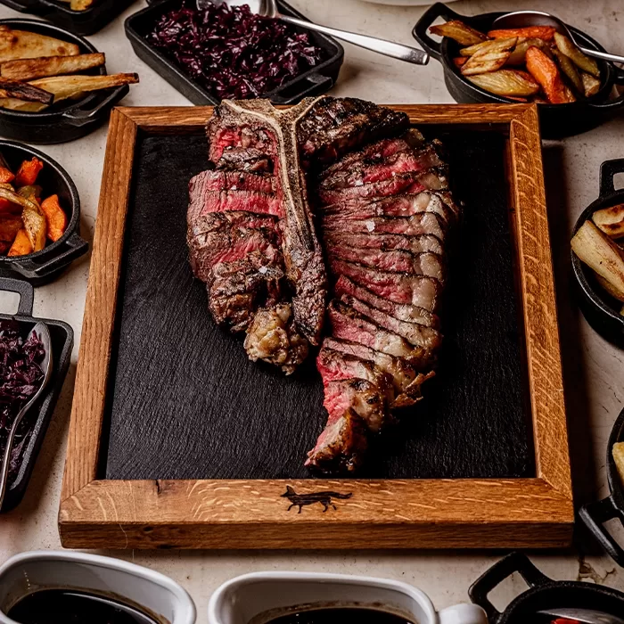 A sliced, medium-rare T-bone steak is served on a wooden board with several sides, including roasted potatoes, carrots, and cooked red cabbage, arranged around it.