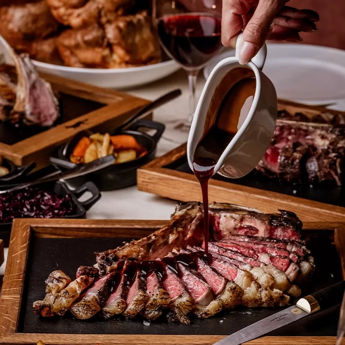 A hand pours rich brown sauce over sliced medium-rare steak on a wooden board, surrounded by side dishes, wine, and other plates of food in a restaurant setting.