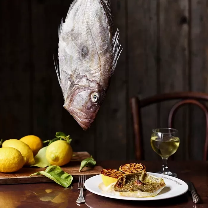A whole raw fish is suspended above a plate of cooked fish with grilled lemon halves, next to a glass of white wine and a cutting board with fresh lemons on a rustic wooden table.