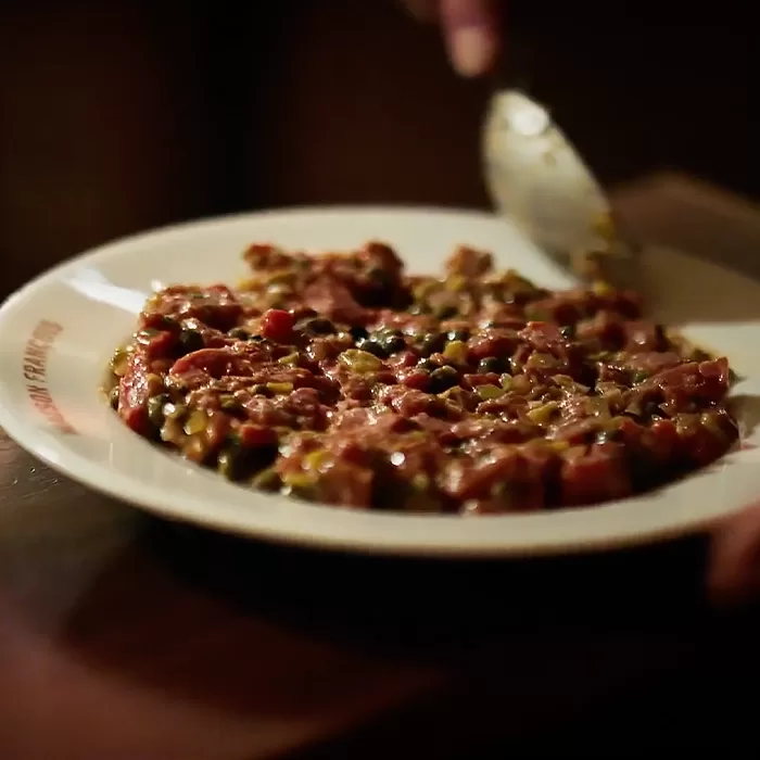 A close-up of a plate with steak tartare, finely chopped raw beef mixed with capers and seasoning, with a hand holding a spoon above the dish.