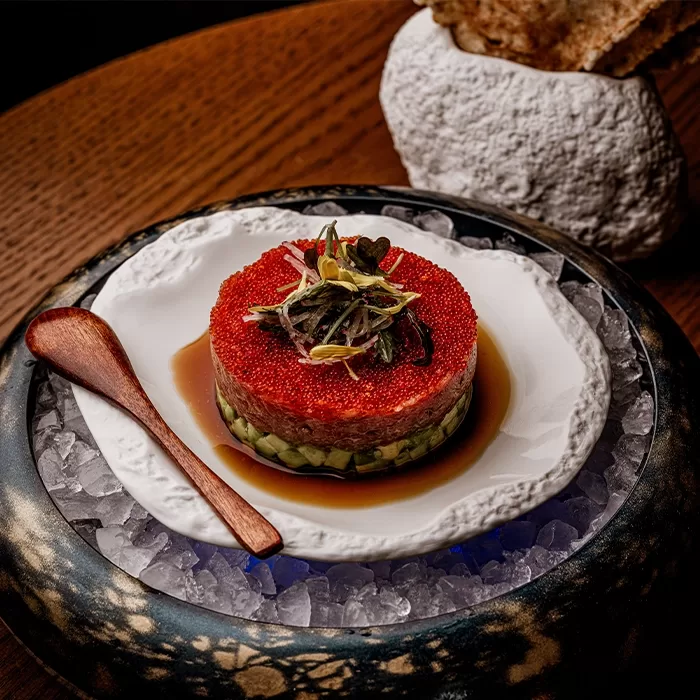 A gourmet dish with layers of diced cucumber, minced tuna, and a topping of bright red fish roe, garnished with microgreens; served on a white plate over ice with a wooden spoon, and crispbread in the background.