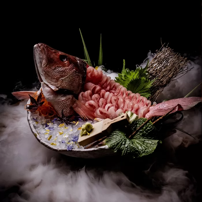 A dramatic sushi platter featuring a whole fish with sashimi slices arranged along its body, surrounded by garnishes, herbs, and misty smoke on a dark background.