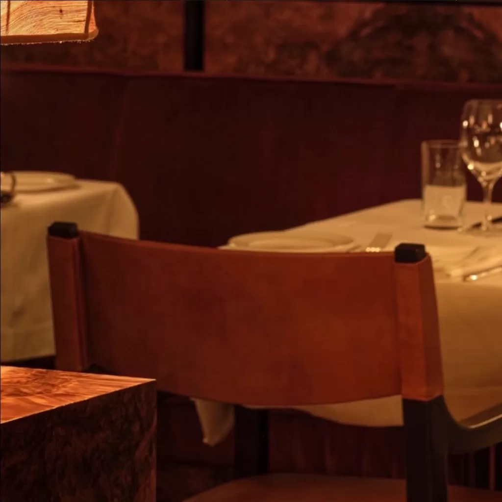 A warmly lit restaurant table set for two with white tablecloth, glassware, and napkins. Brown leather chairs and a cozy, intimate ambiance are visible.