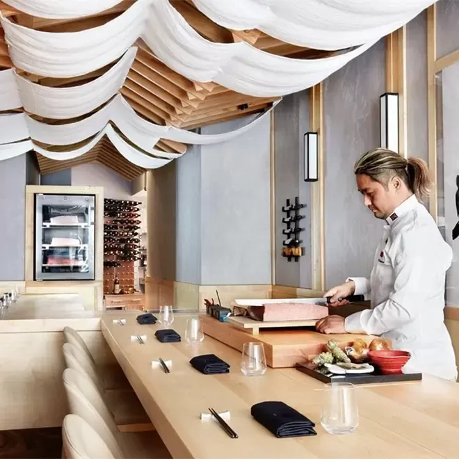 At Marru restaurant, a chef meticulously prepares dishes at a modern sushi counter with wooden elements and a draped ceiling. Empty chairs line the bar, while a wine display graces the background, creating an inviting ambiance.
