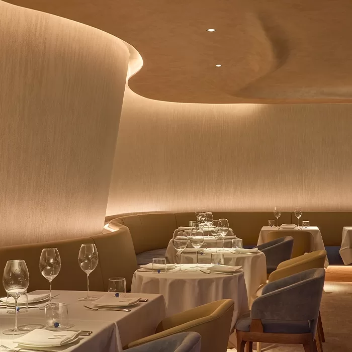 Elegant, softly lit restaurant interior with curved beige walls, round tables covered in white cloths, neatly set with wine glasses and plates, and cushioned chairs. The atmosphere is modern and inviting.
