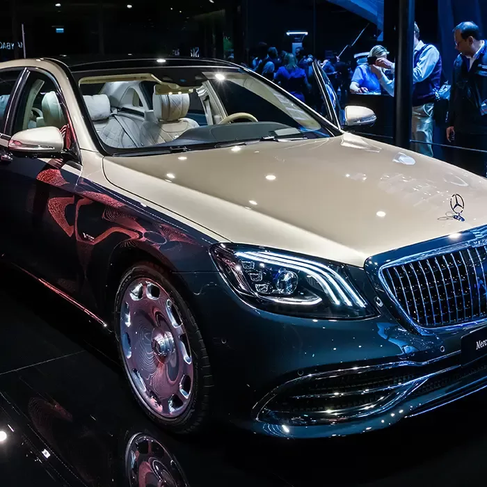 A luxury Mercedes-Benz Maybach sedan is displayed at an auto show, with a two-tone exterior, chrome accents, and a plush white leather interior, attracting attention from onlookers in the background.
