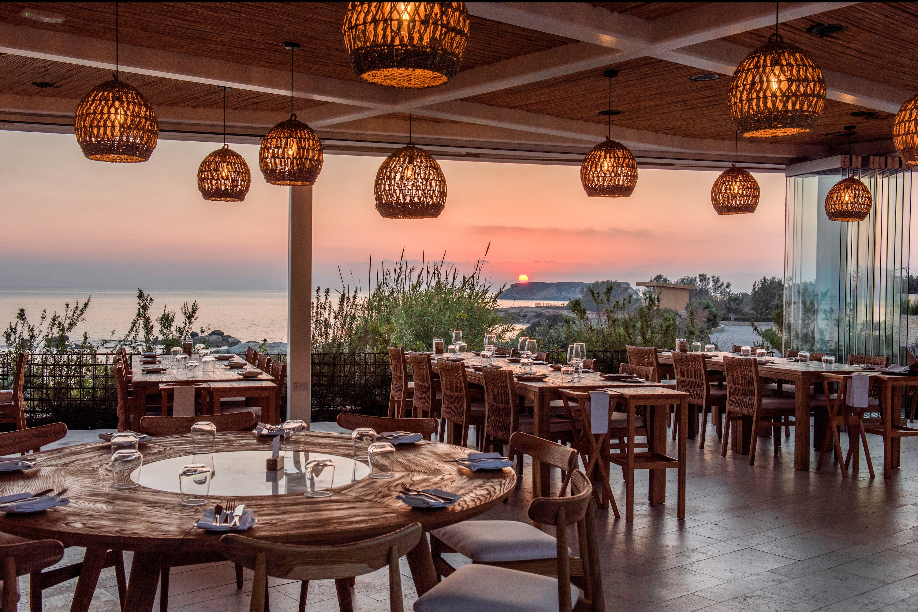 Outside restaurant seating area with variety of round and rectangle wooden tables and cushioned chairs with sunset sea view.