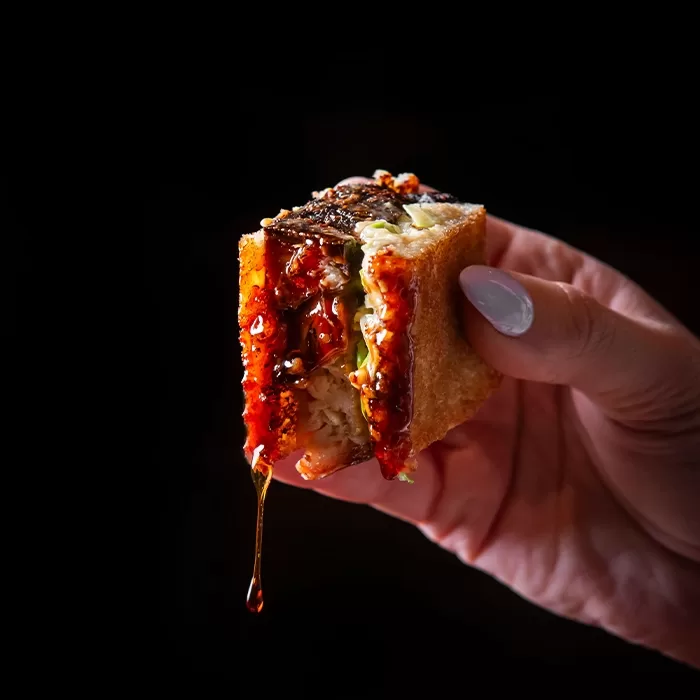 A hand with manicured nails holds a piece of sushi with a golden-brown crispy exterior, spicy sauce, rice, and fish, with sauce dripping from the edge against a black background.