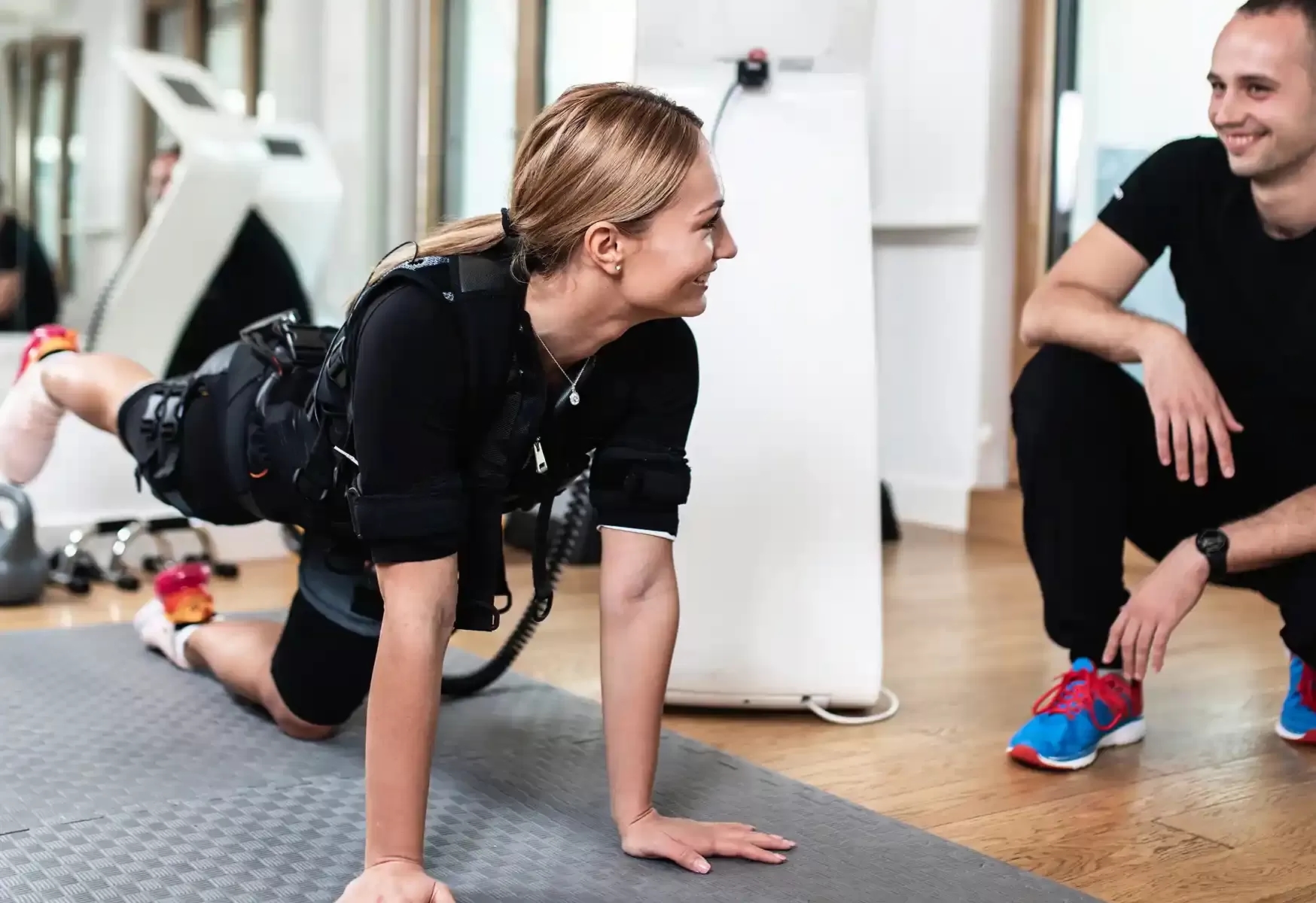 Women on training mats in gym on the floor using a EMS (electro-muscle stimulation) smiling at Personal Trainer