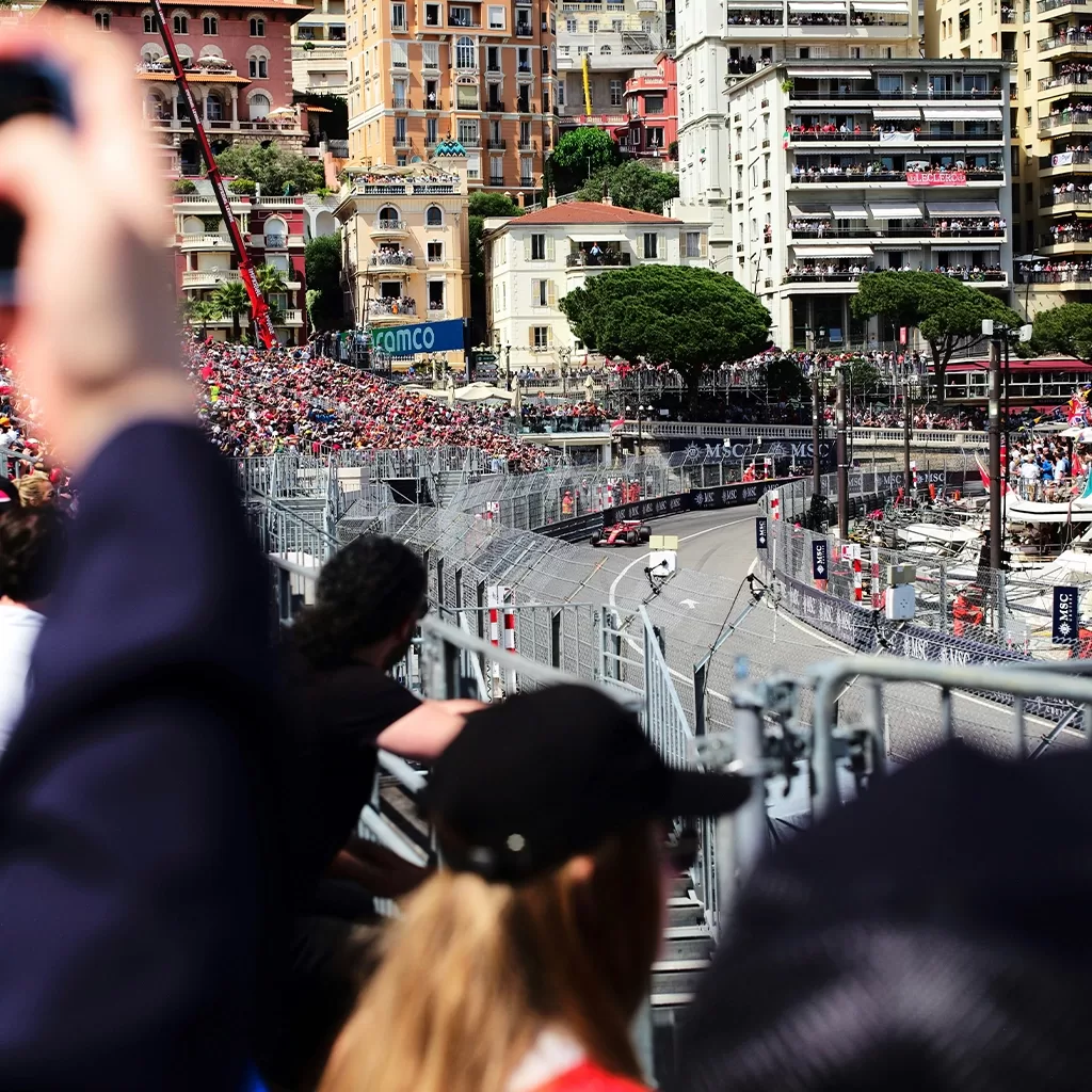 A crowd watches a Formula 1 race as cars speed around a curve on a city street circuit, with tall buildings and yachts in the background. Some spectators in the foreground hold up phones to take photos.