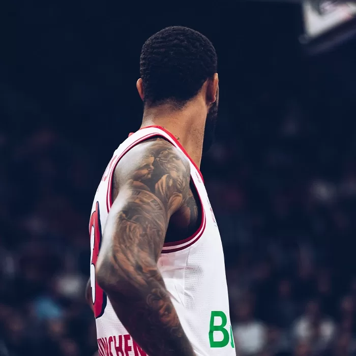 A basketball player wearing a white and red jersey with the number 0 stands with his back turned. His arms are covered in tattoos, and the background shows a blurred crowd.