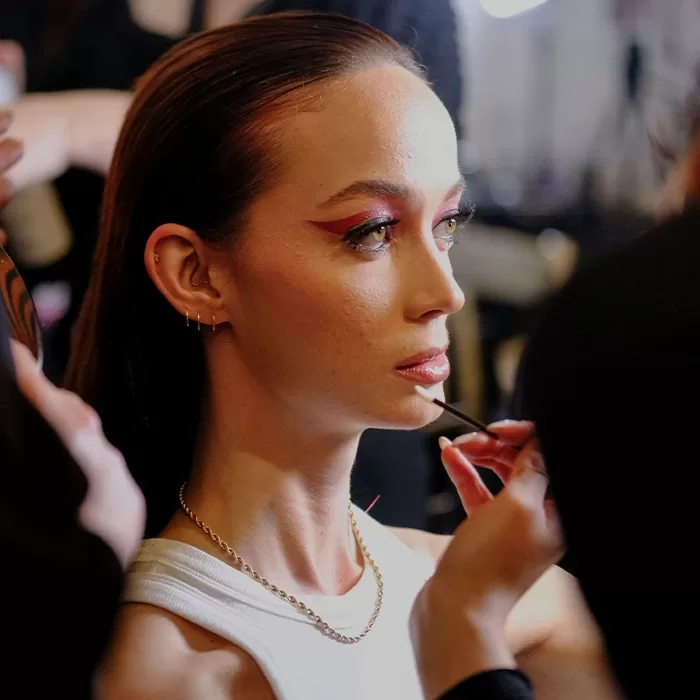 A person with slicked-back hair receives a makeup touch-up, with bold red eyeliner and natural lip color, while wearing a white top and a chain necklace. Multiple hands apply makeup on their lips and face.