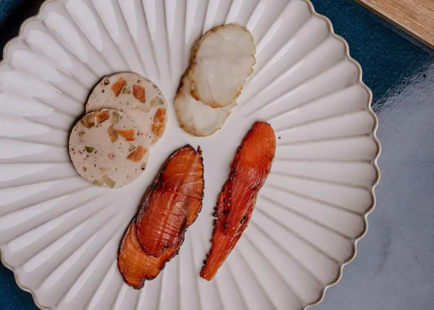 A white plate with a scalloped edge displays gourmet food: sliced terrine with visible vegetables, a piece of white fish, and two pieces of seared salmon. The plate rests on a dark blue surface.