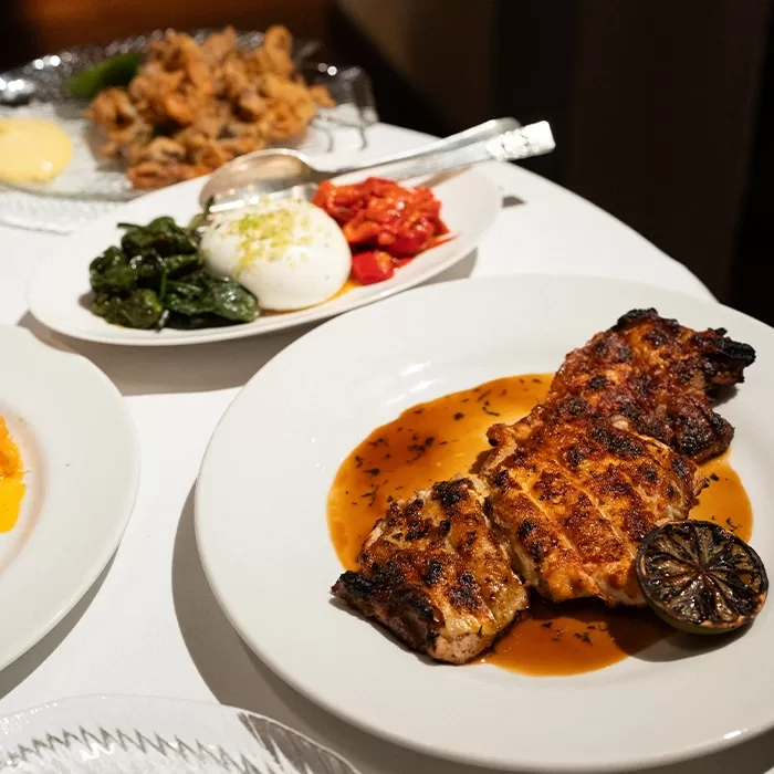Grilled chicken with sauce and a charred lime on a white plate, alongside plates of burrata with vegetables and a fried dish, all served on a white tablecloth.