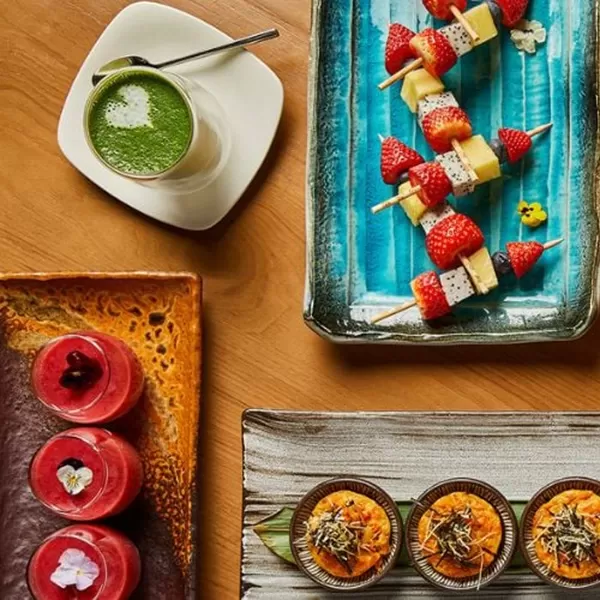 Top view of a table with colorful dishes: a creamy green drink, fruit and cheese skewers, three small bowls with orange-topped dishes, and a rectangular plate with pink desserts decorated with flowers at Nobu, London.
