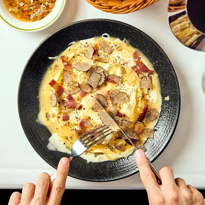 A person uses a knife and fork to cut into a creamy pasta dish topped with shaved truffles, bacon pieces, and black pepper, served on a black plate on a white tablecloth.