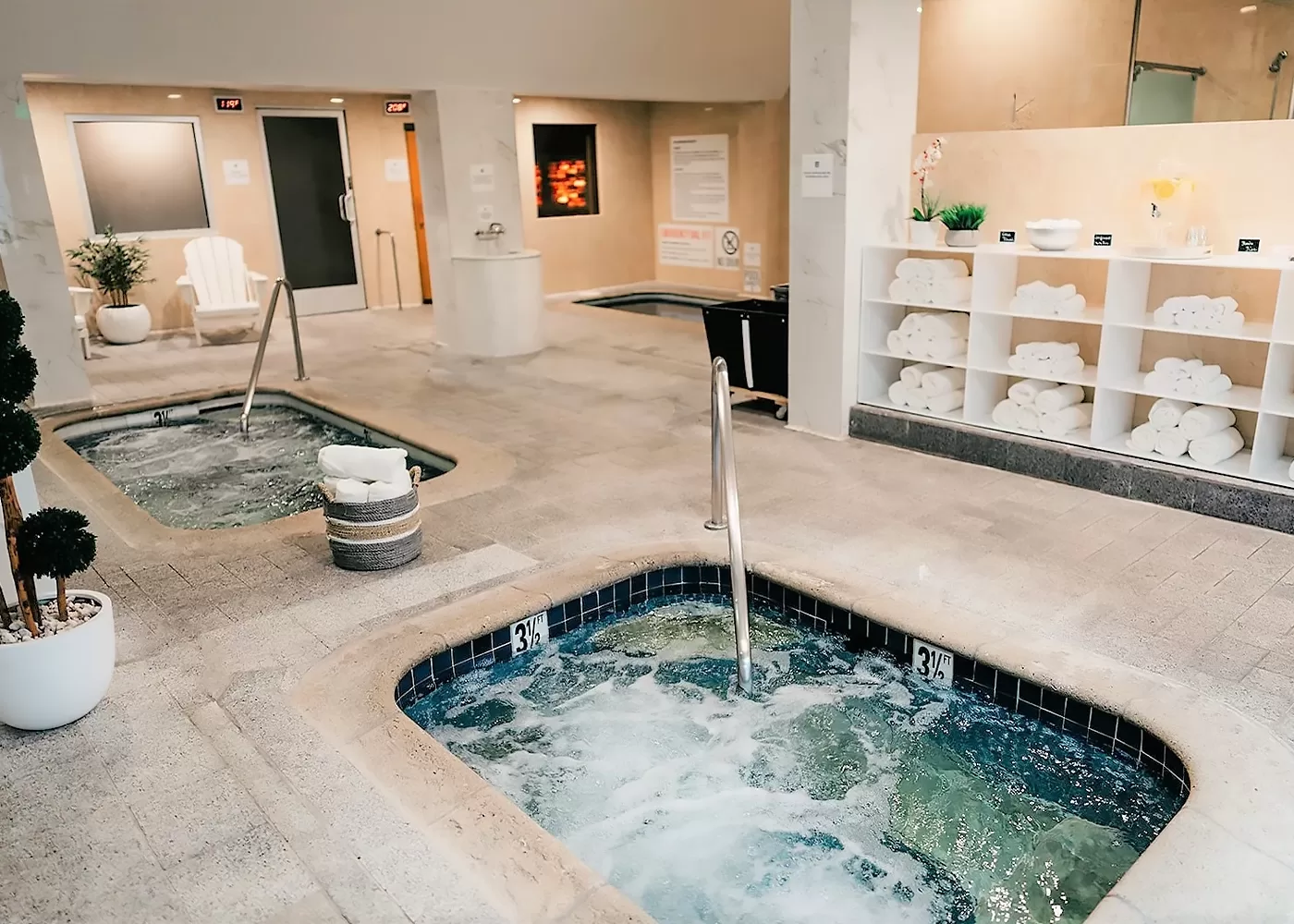 Spacious indoor spa area with bubbling hot tubs, rolled white towels on open shelves, potted plants, and relaxing seating. Warm lighting creates a tranquil, inviting atmosphere.