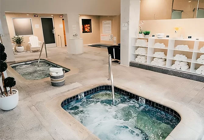 Indoor spa area with two bubbling hot tubs, shelves filled with rolled white towels, white chairs, potted plants, and warm lighting creating a relaxing atmosphere.