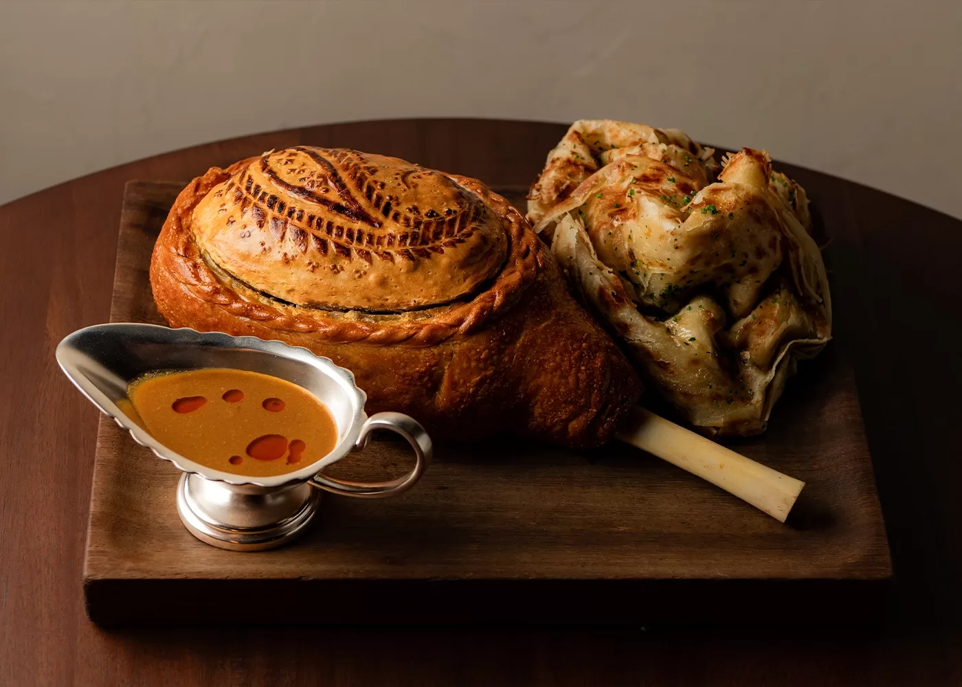 A wooden board with a baked meat pie on a bone, patterned crust on top, flatbread folded beside it, and a silver gravy boat filled with orange sauce, all placed on a dark brown table.