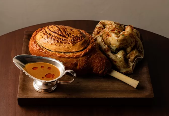 A wooden board displays a baked leg of lamb wrapped in golden brown pastry, a stack of folded flatbread, and a metal gravy boat with reddish-brown sauce, garnished with drops of oil.