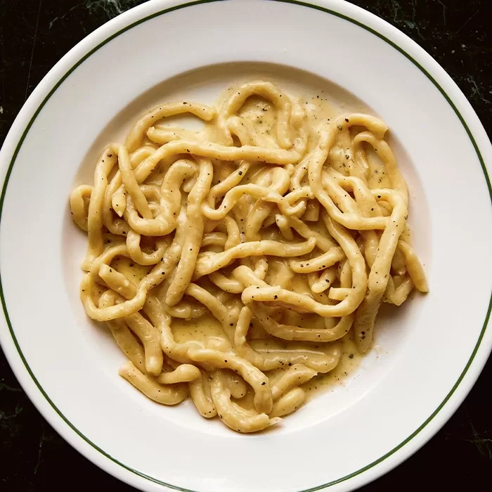 A plate of thick, hand-rolled pasta coated in a creamy, pepper-speckled sauce, served on a white dish with a green rim, on a dark surface.