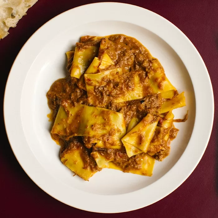 A white plate on a dark red surface holds wide, flat pasta sheets covered in a thick, rich brown meat sauce.