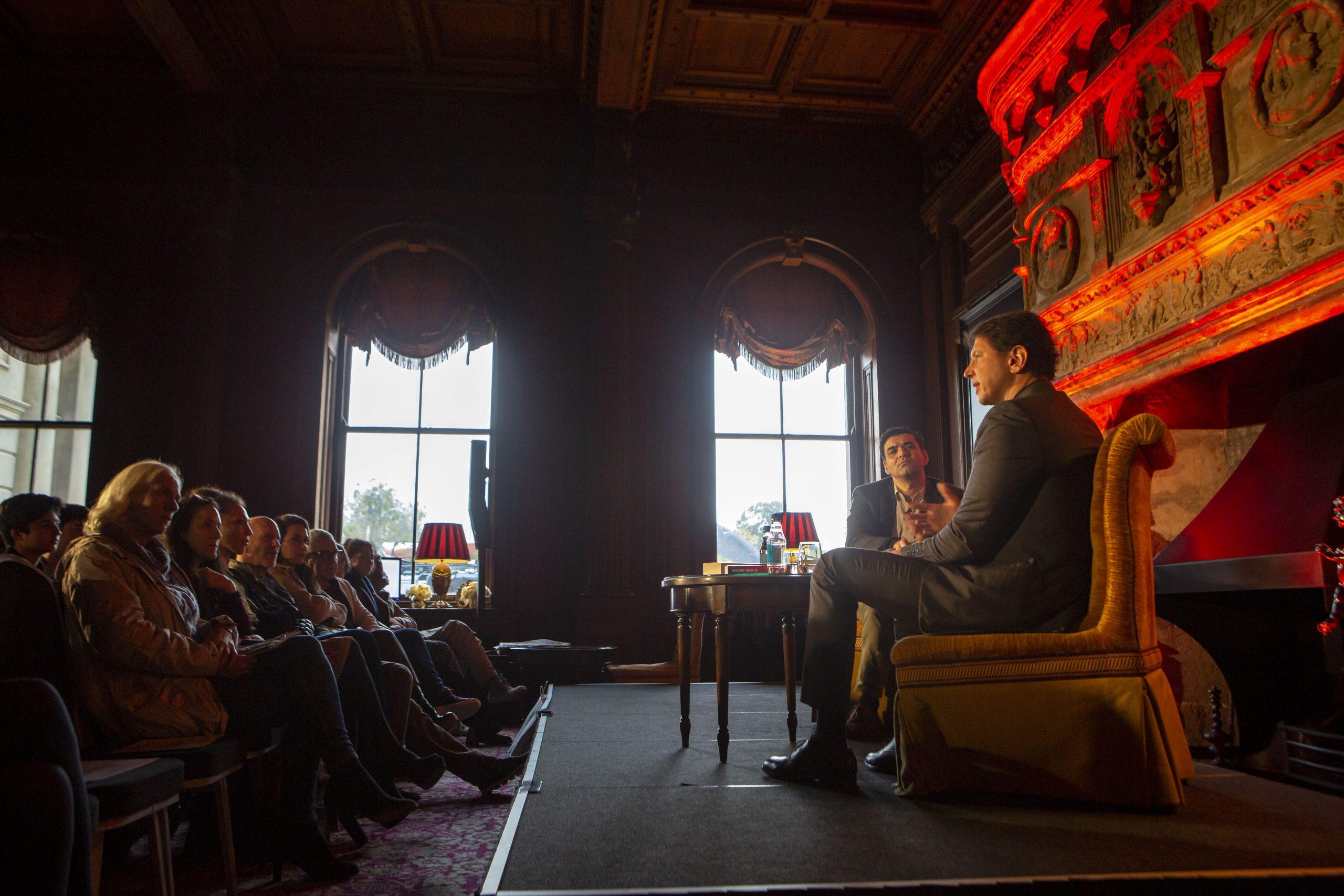 Audience on chairs facing two men on a stage sat on luxury chairs in front of red grand fireplace doing a panel discussion at Cliveden.