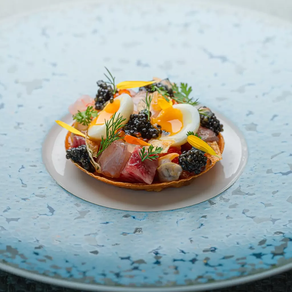 A gourmet tart topped with diced fish, a soft-boiled egg, black caviar, edible flowers, and fresh herbs, served on a speckled blue and white plate.