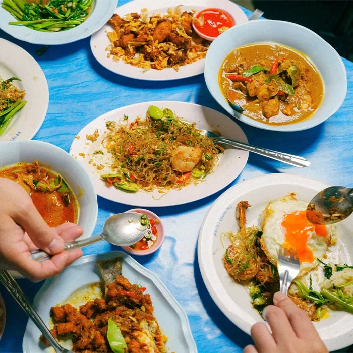A table set with various Thai dishes on white plates, including stir-fried noodles with shrimp, curry, vegetables, fried fish, and a plate with a runny fried egg, with hands reaching in to serve food.