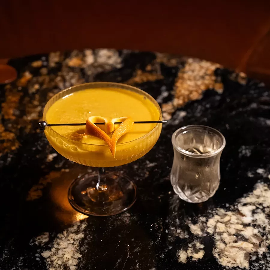 A yellow cocktail garnished with a twisted orange peel on a pick sits in a coupe glass on a dark, marbled table beside a small glass of clear liquid.