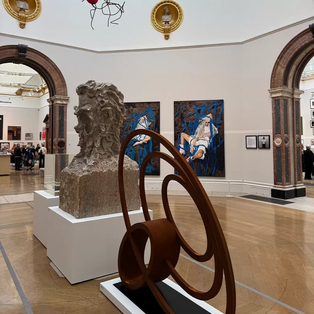 A museum gallery features a stone bust and a circular metal sculpture in the foreground, with paintings and framed photos on white walls and visitors viewing art in the background.