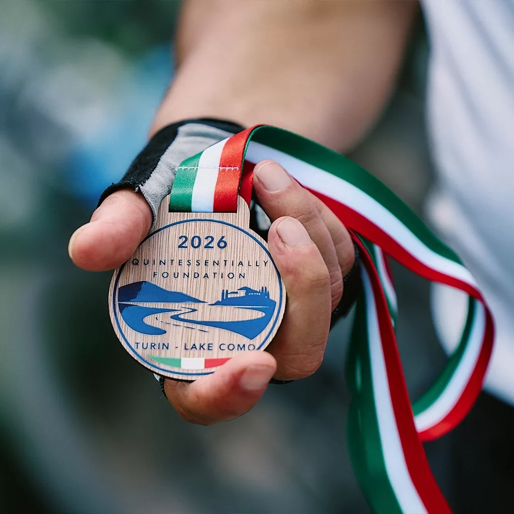 A hand holds a wooden medal with an Italian flag ribbon. The medal reads 2026 Quintessentially Foundation Turin-Lake Como and features a stylized river and mountain design.