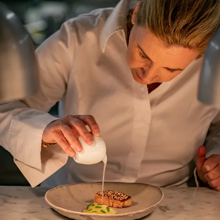 A chef in a white coat carefully pours a white sauce from a small pitcher onto a plated dish, focusing on presentation in a professional kitchen setting.