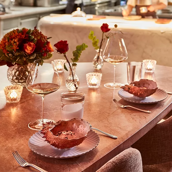 A warmly lit restaurant table set with two plates holding seafood shells, wine glasses, a glass of water, flowers in vases, and lit candles, creating an elegant and romantic dining atmosphere.