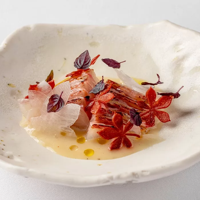 A white ceramic bowl with artfully arranged sashimi, garnished with red flower-shaped herbs, small purple leaves, and a light sauce with droplets of oil. The presentation is delicate and minimalist.