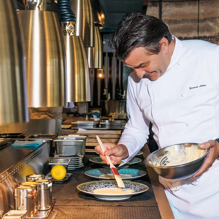 A chef in a white coat carefully brushes sauce onto a plate in a modern kitchen, holding a mixing bowl in one hand and focusing on plating the dish.