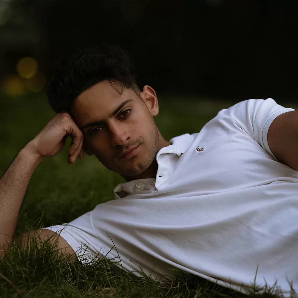 A young man in a white polo shirt lies on grass, resting his head on one hand and looking at the camera with a relaxed expression. The background is dark and blurry, emphasizing the subject.