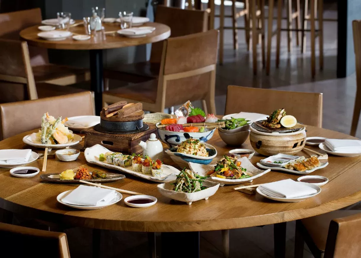 A round wooden table is set with various Japanese dishes, including sushi, tempura, and edamame, on ceramic plates. There are empty plates and chopsticks ready for diners, with chairs surrounding the table in a cosy restaurant setting at Roka, London.