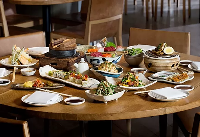 A round wooden table is set with various Japanese dishes, including sushi, tempura, and edamame, on ceramic plates. There are empty plates and chopsticks ready for diners, with chairs surrounding the table in a cosy restaurant setting at Roka, London.