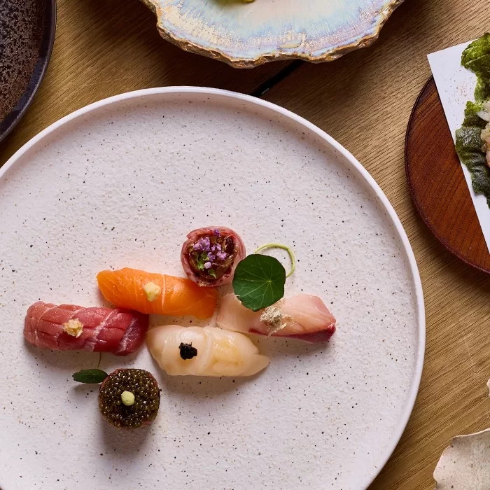A white plate with assorted sushi pieces, including tuna, salmon, scallop, uni, and garnishes, arranged artfully. Other dishes and a piece of seaweed are partially visible around the main plate.