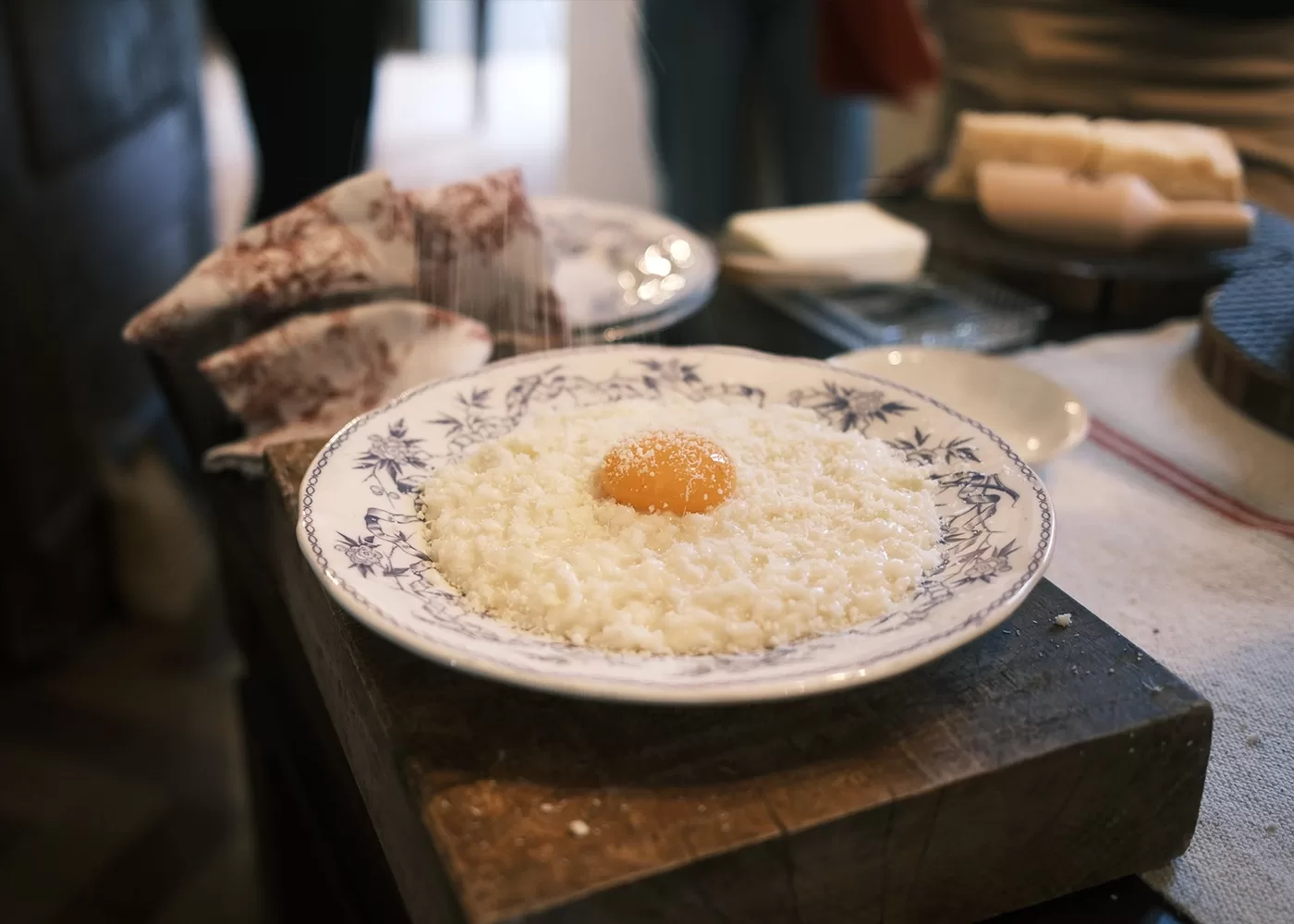A decorative plate with white rice topped with a raw egg yolk sits on a wooden surface, surrounded by kitchen items and blurred people in the background.