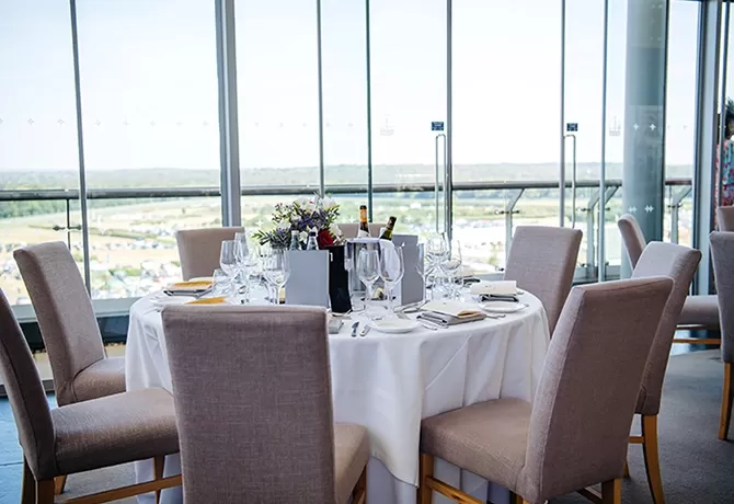 Ascot Royal Enclosure’s round table and chairs with fine dining set up by a glass window and view of the racecourse at the Panoramic Restaurant