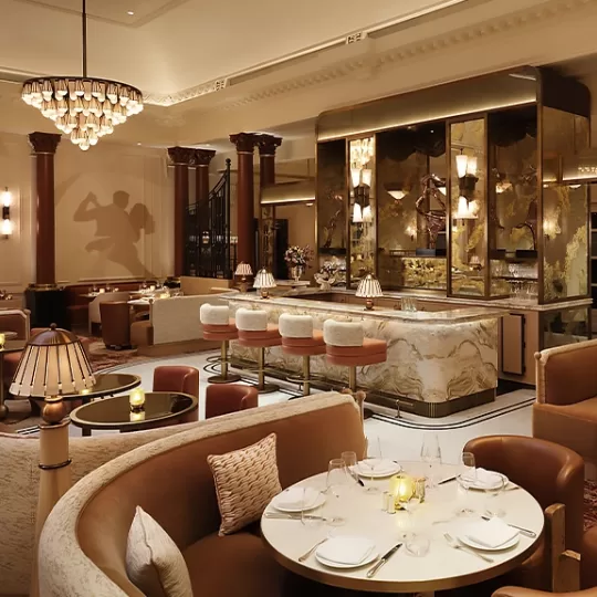 Elegant restaurant interior with round tables set for dining, plush curved booths, marble-topped bar with high stools, warm lighting from chandeliers and lamps, and gold accents throughout the space.