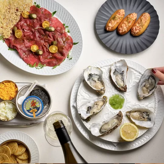 A table set with oysters on a white platter, a hand reaching for one, beef carpaccio with capers and yellow tomatoes, lobster toasts on a black plate, a tin of caviar with accompaniments, lemon, and a bottle being poured.